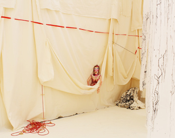 Figure in red dress partially emerging from draped cream-coloured fabric in an installation space, surrounded by hanging sheets and red rope.