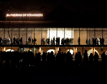 exterior view of the gallery at night with 'Gathering of strangers' written in lights with silhouettes of visitors in the gallery.