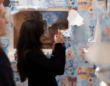 A woman is photographing an artwork inside a wallpapered space.