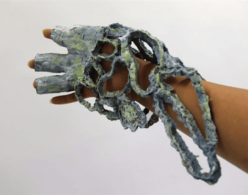 A blue and green papier-mâché glove on an outstretched hand against a white background.