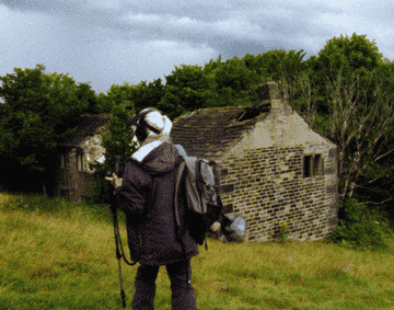 A person wearing headphones stands in front of an old cottage in a green field