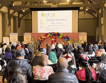 A group of people are sat listening to Qaisra Shahraz speak in front of a presentation.