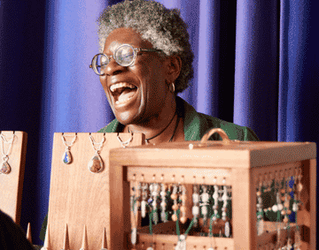 An Black craftswoman laughing behind a jewellery display in front of a purple curtain