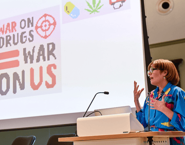 A presenter stands in front a large screen displaying various cross-stitch patterns. 
