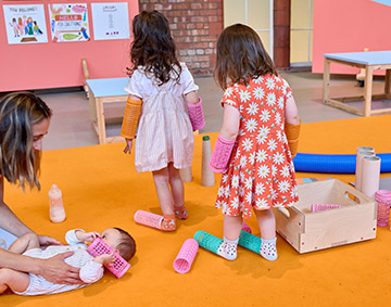 Two children are playing in the gallery with orange flooring - there are art materials - in the gallery's studio space.