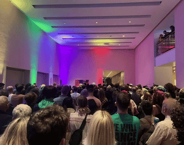 A large crowd of people inside a white walled gallery space, decorated with colourful lights