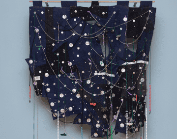 A textile piece made out of navy healthcare uniforms, with talismans and beads hanging from the piece.